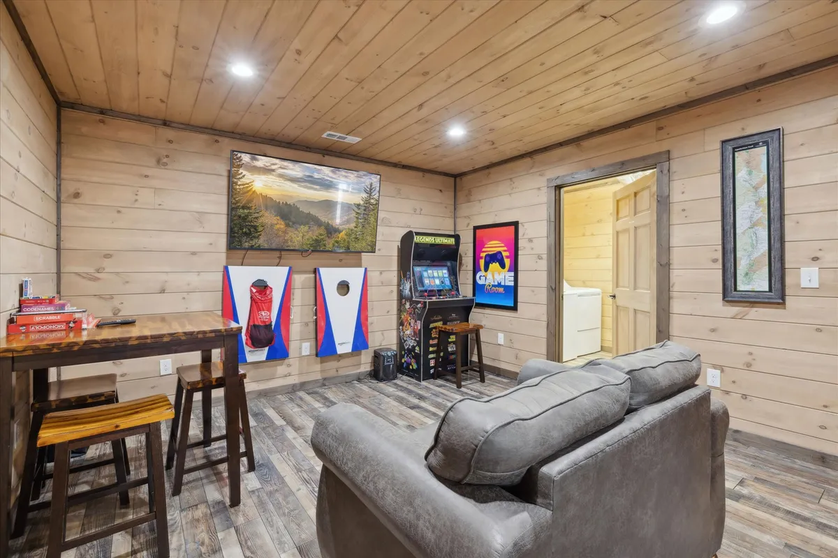 A vibrant interior game room featuring a wooden table, gray sofa, an arcade game machine, wall art, and a bean bag toss game. The setup invites fun, friendly competition, and family gatherings.