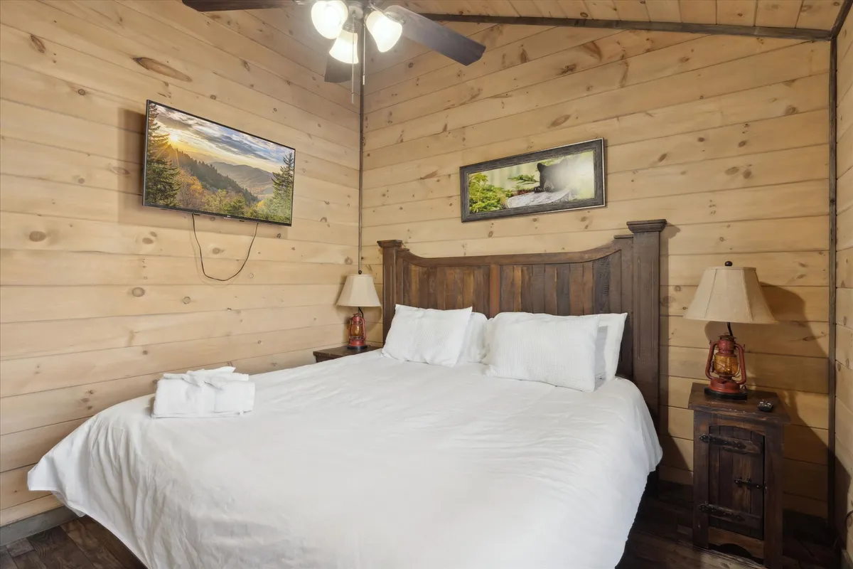 A serene bedroom with a bed dressed in white linens and a sturdy wooden headboard. Soft lighting from two bedside lamps enhances the tranquil atmosphere, while a wall-mounted TV and framed nature artwork add modern comforts and a touch of the outdoors. The space feels restful and private, perfect for unwinding after a day of adventure.