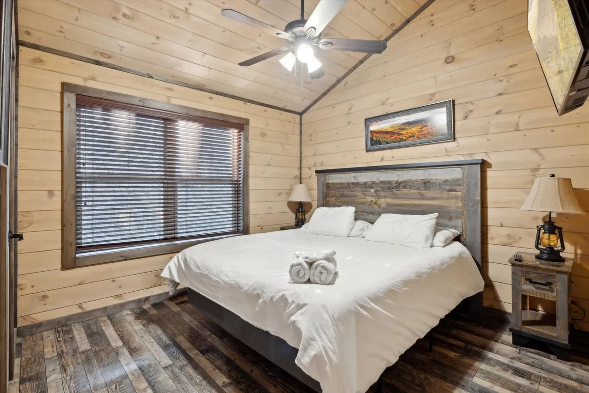 A welcoming bedroom centered around a king-size bed with soft, fresh linens. The cozy cabin décor features wooden ceilings, warm-toned walls, and nightstands with glowing lamps. A decorative painting hangs above the bed, and a window with blinds allows natural light to stream in, creating an inviting and tranquil atmosphere ideal for couples or solo travelers seeking rest and romance.