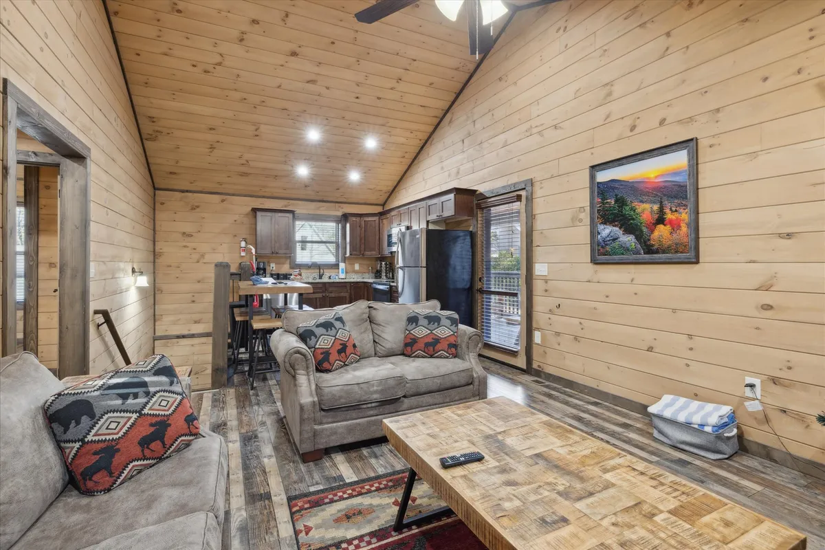 A relaxed open-concept living and kitchen area with a comfortable sofa adorned with patterned cushions, a wooden coffee table, and a view of the kitchen in the background. Landscape artwork and windows create a connection to the outdoors, while the space feels both homey and versatile for different group activities.