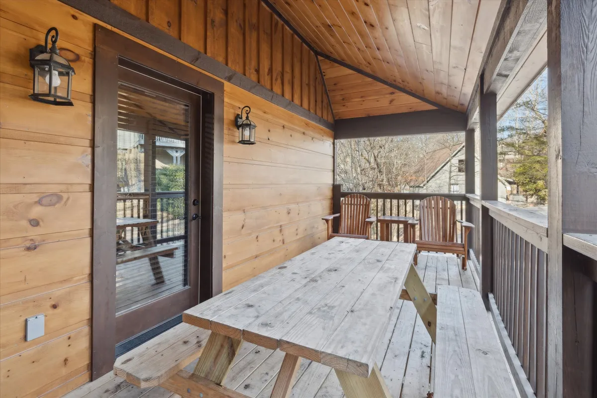 A cozy porch scene with a wooden picnic table, two Adirondack chairs, lantern-style light fixtures, and a door leading inside. The porch offers a warm, inviting space to enjoy morning coffee or evening drinks while taking in views of the Smoky Mountains.