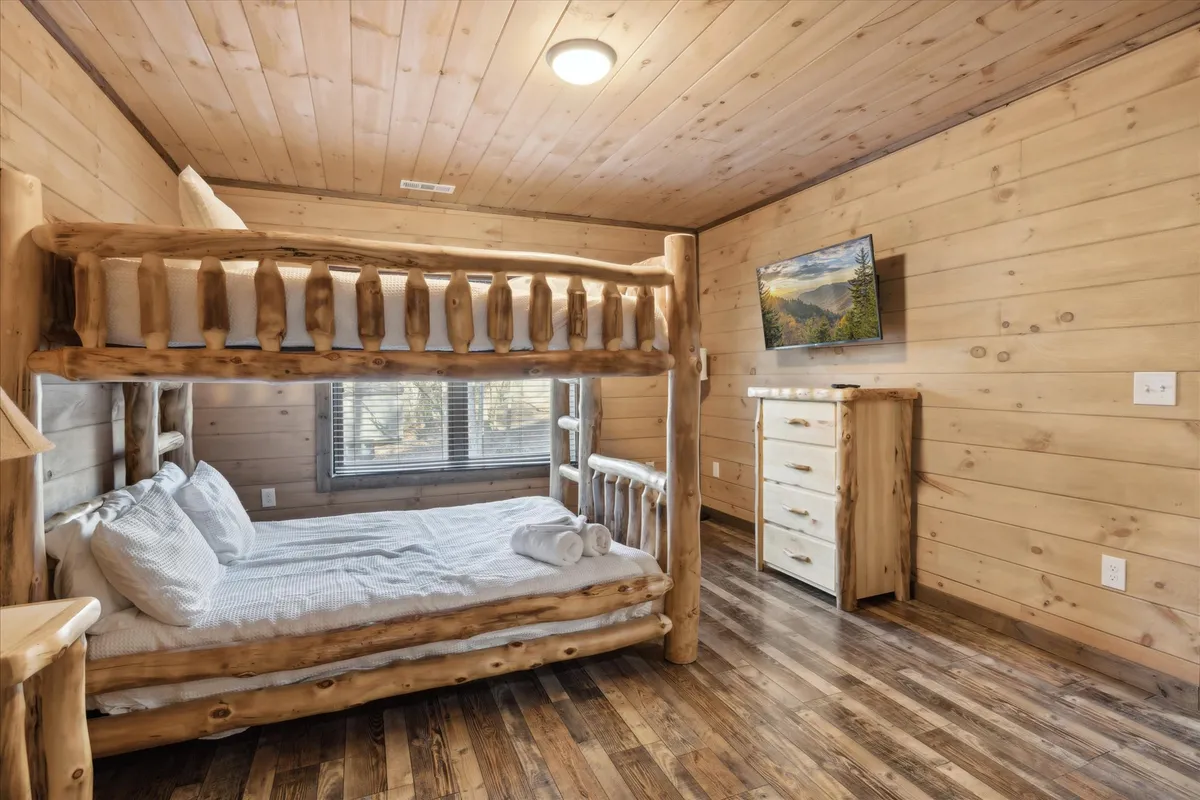 A bunk bed setup with a wooden dresser, wall-mounted TV, and windows, this inviting room suggests a cozy retreat for groups or families to rest and recharge after a day of Smoky Mountain adventures.