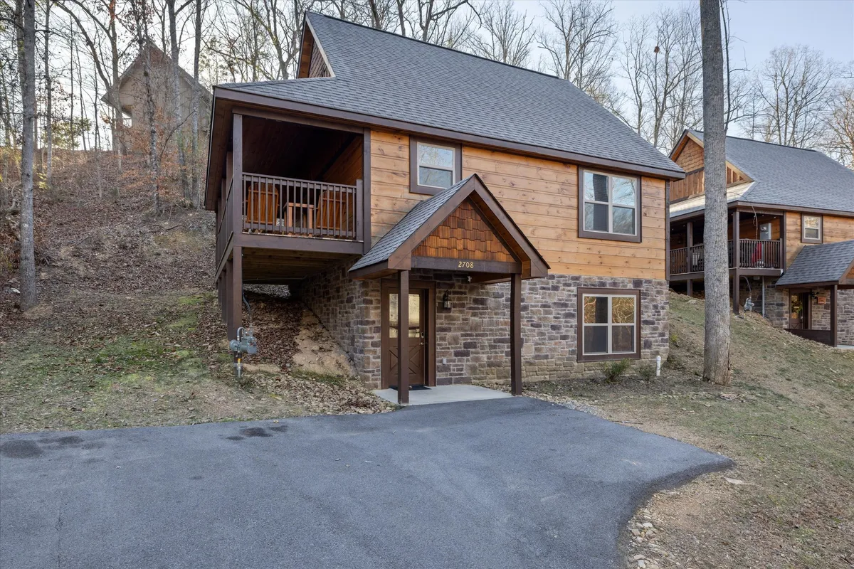 This image shows the welcoming exterior of the cabin, including the wooden facade, stone foundation, balcony, and entrance pathway. The surrounding natural landscape hints at the cabin's tranquil setting, with space for outdoor relaxation and gatherings on the porch and balcony.