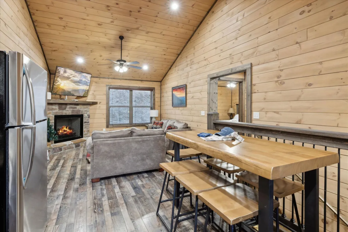 This image highlights a spacious open-plan area featuring a crackling fireplace, a wooden dining table set for meals, a plush sofa, and large windows that let in natural light. The setup invites families or friends to share meals, relax by the fire, or simply enjoy the mountain views from indoors.