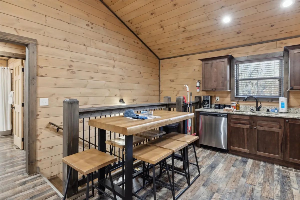 A spacious dining area with a large wooden dining table, modern kitchen appliances visible in the background, exposed wooden beams, and bar stools. The setting is ideal for family meals, casual gatherings, or sharing stories over dinner.